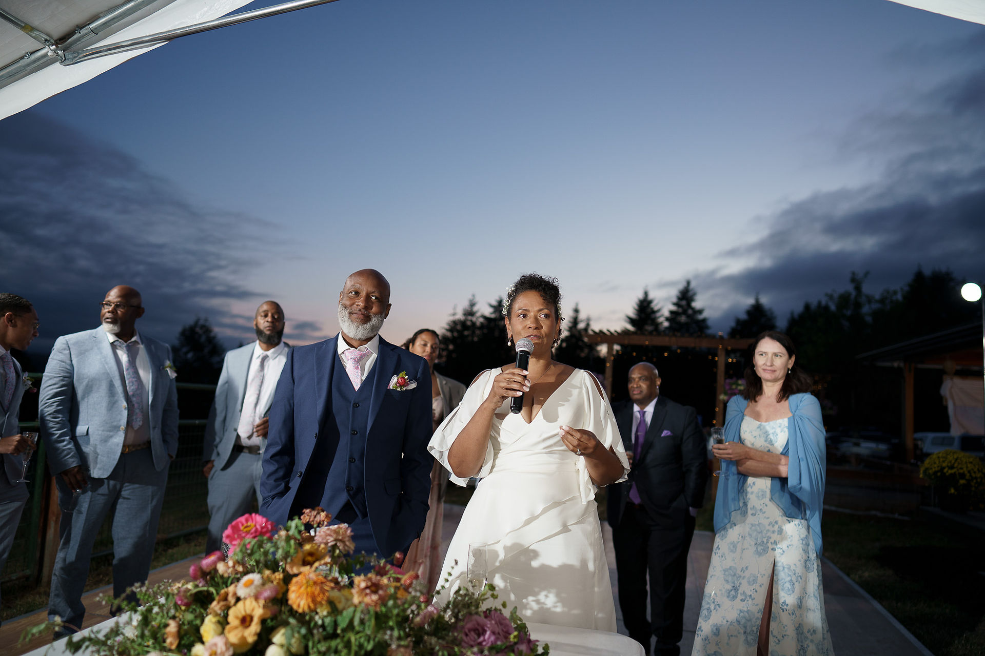 Bride speaking into microphone at outdoor wedding reception at dusk.