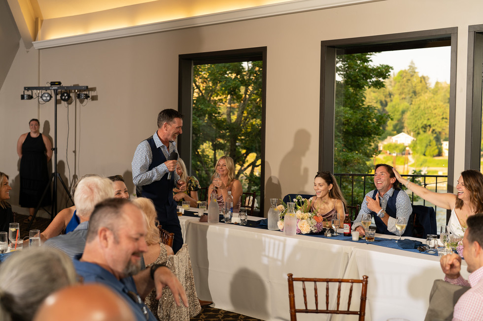 Man giving speech at wedding reception to seated, smiling guests.