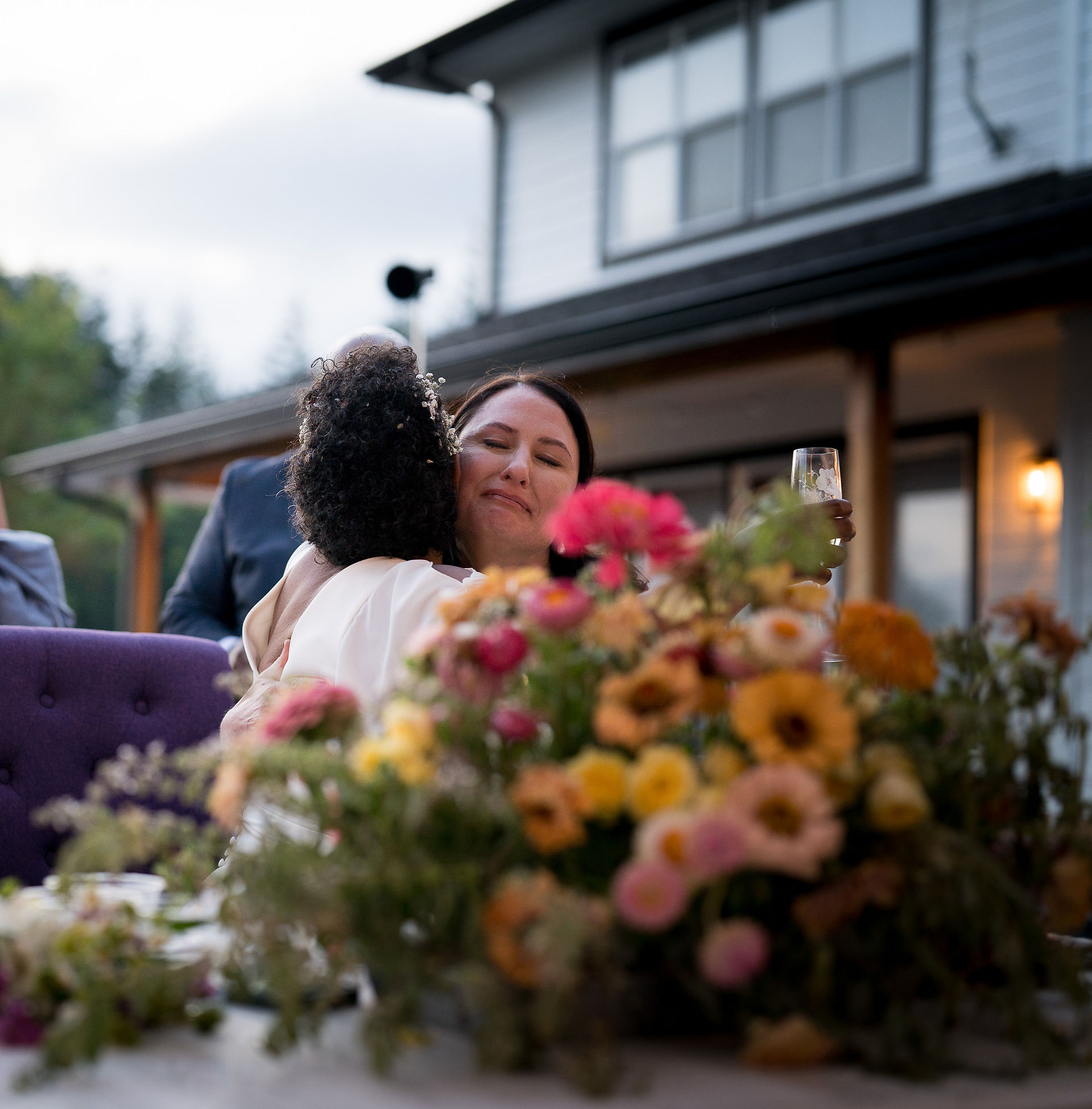 Two women hugging warmly at outdoor wedding with vibrant flowers.