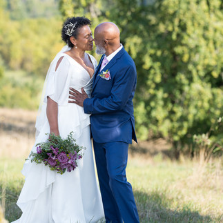 Black bride in white dress and groom in blue suit embrace outdoors.