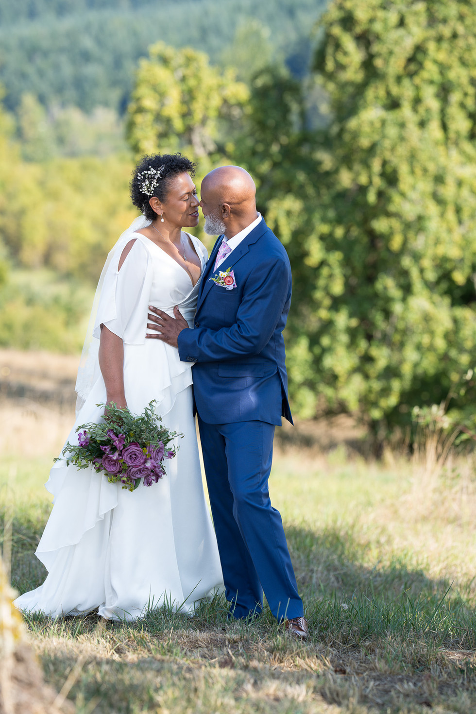 Black bride in white dress and groom in blue suit embrace outdoors.
