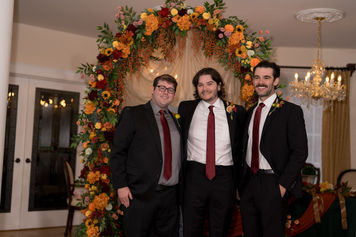Three groomsmen in suits with red ties before fall floral wedding arch.