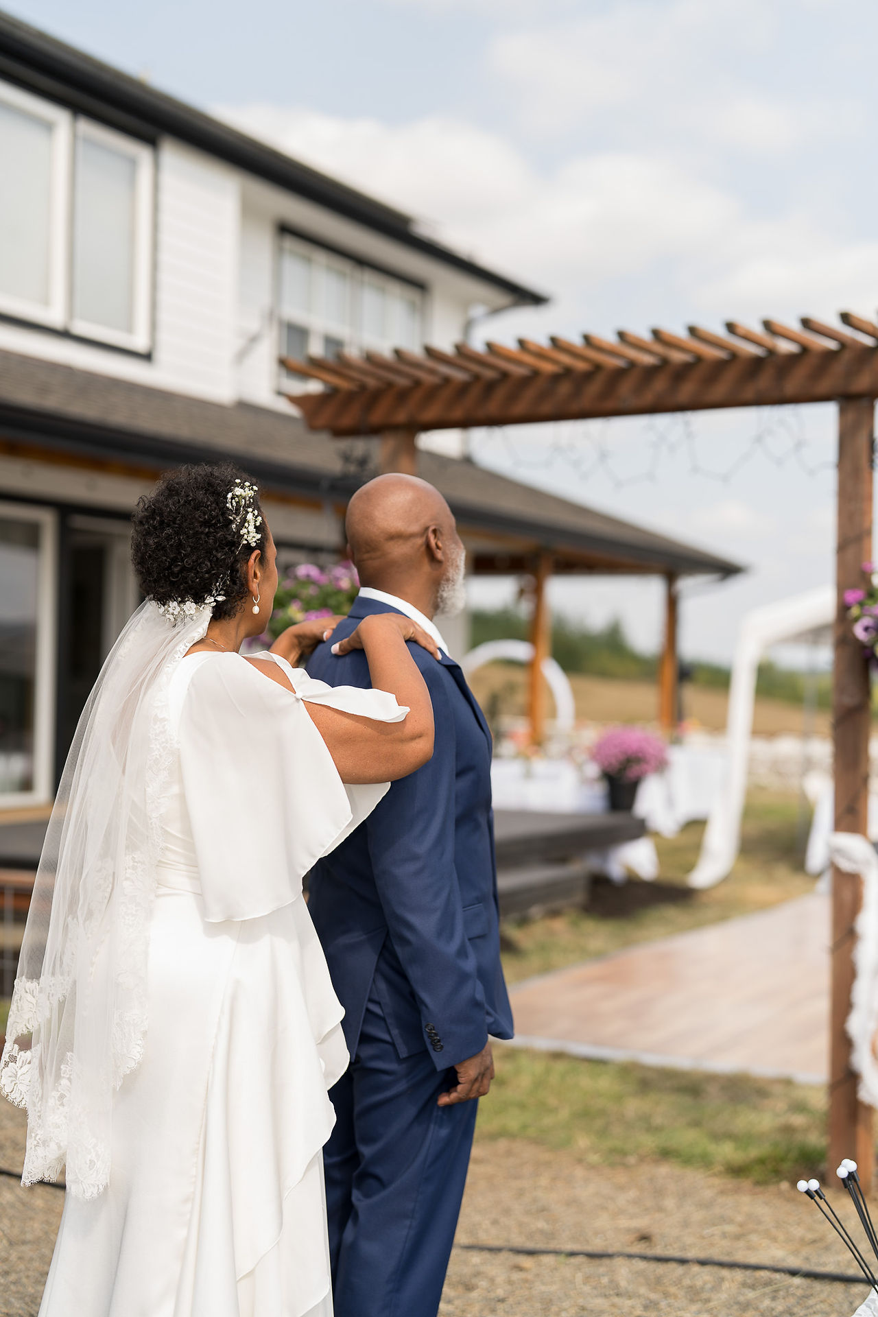 Bride in white dress touches groom's shoulders at intimate farm wedding.