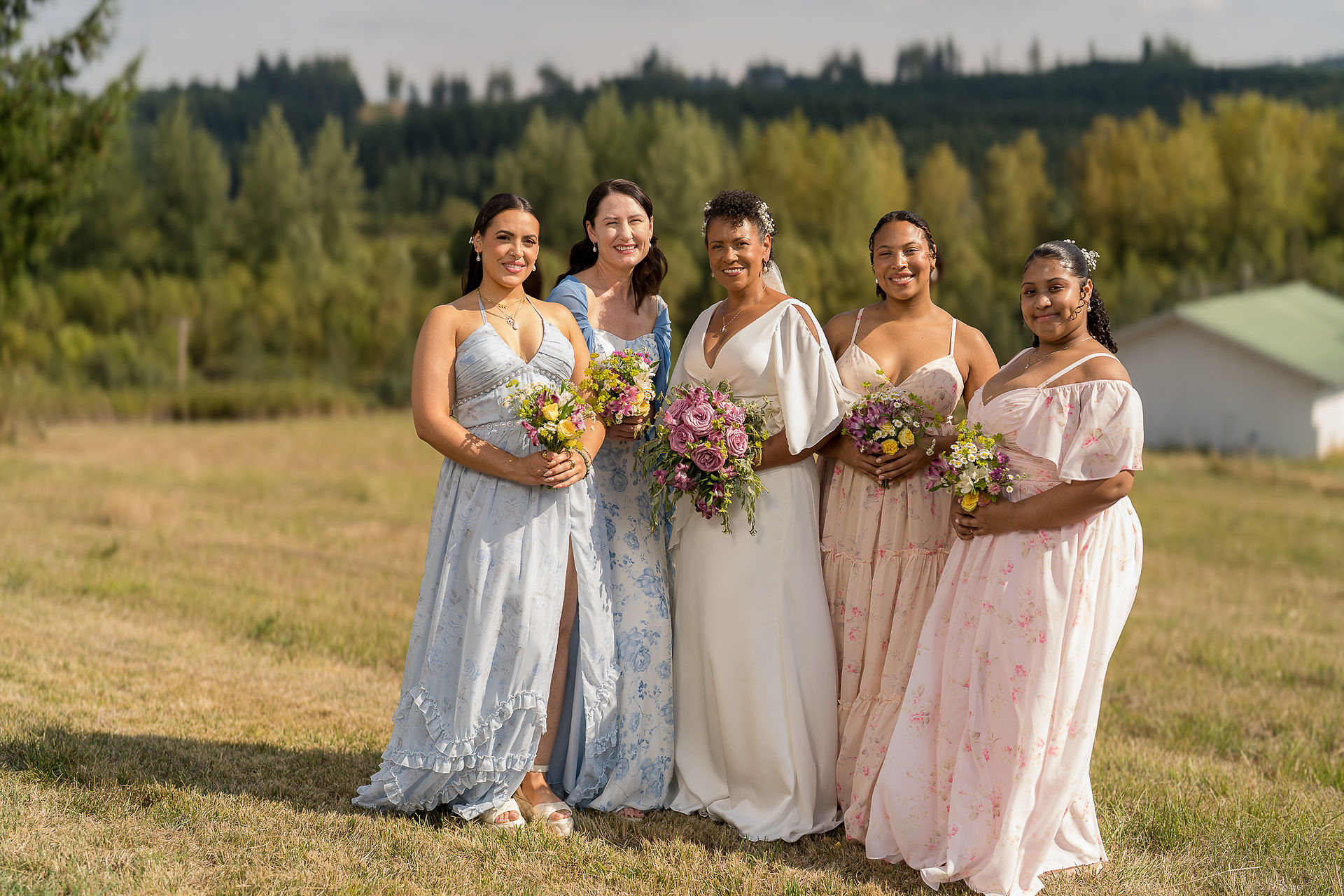 Smiling bride and bridesmaids hold bouquets in a sunny outdoor field.