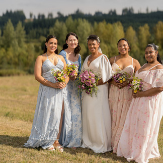 Smiling bride and bridesmaids hold bouquets in a sunny outdoor field.