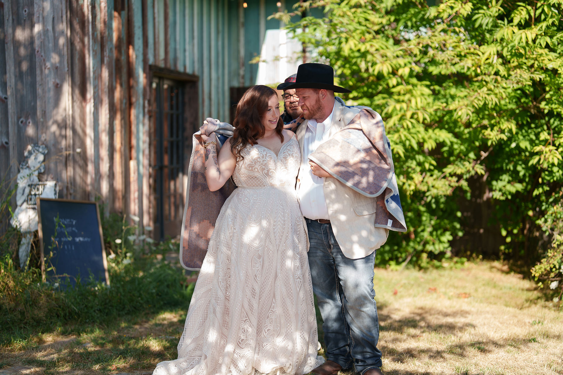 Bride and groom holding blankets, standing outdoors by rustic barn.