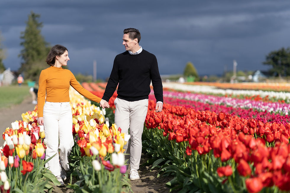 Woman in white dress with straw hat smiles, seated by a canal lined with vibrant red tulips. Floral gardens in the background.