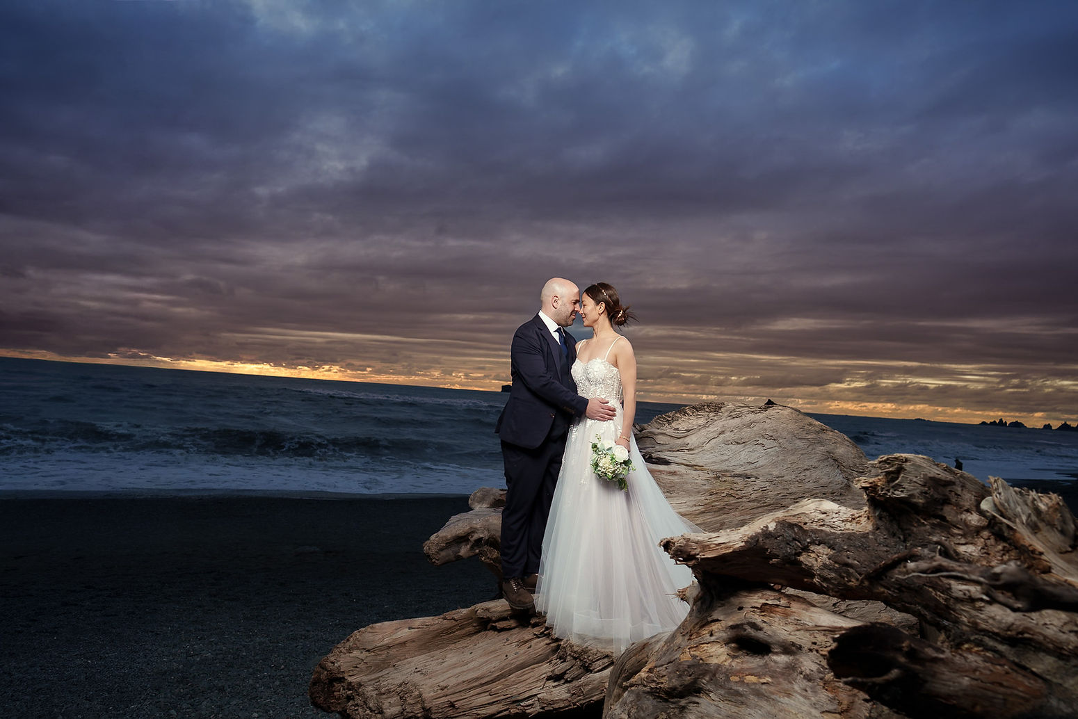 Eloping couple on driftwood at a dramatic PNW beach sunset.