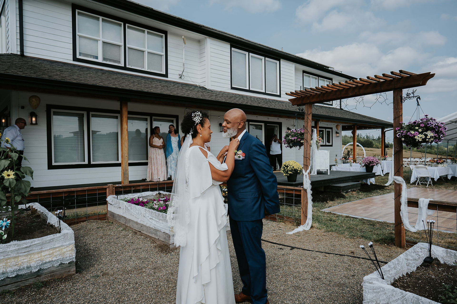 Bride adjusts groom's tie outside white farmhouse on their wedding day.
