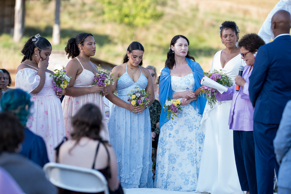 Four women in formal dresses with bouquets at outdoor wedding.