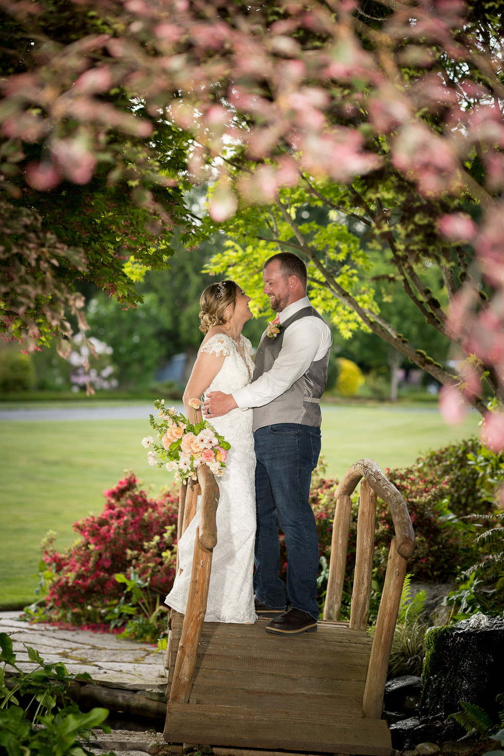 Romantic wedding couple embracing on rustic bridge under pink blossoms.