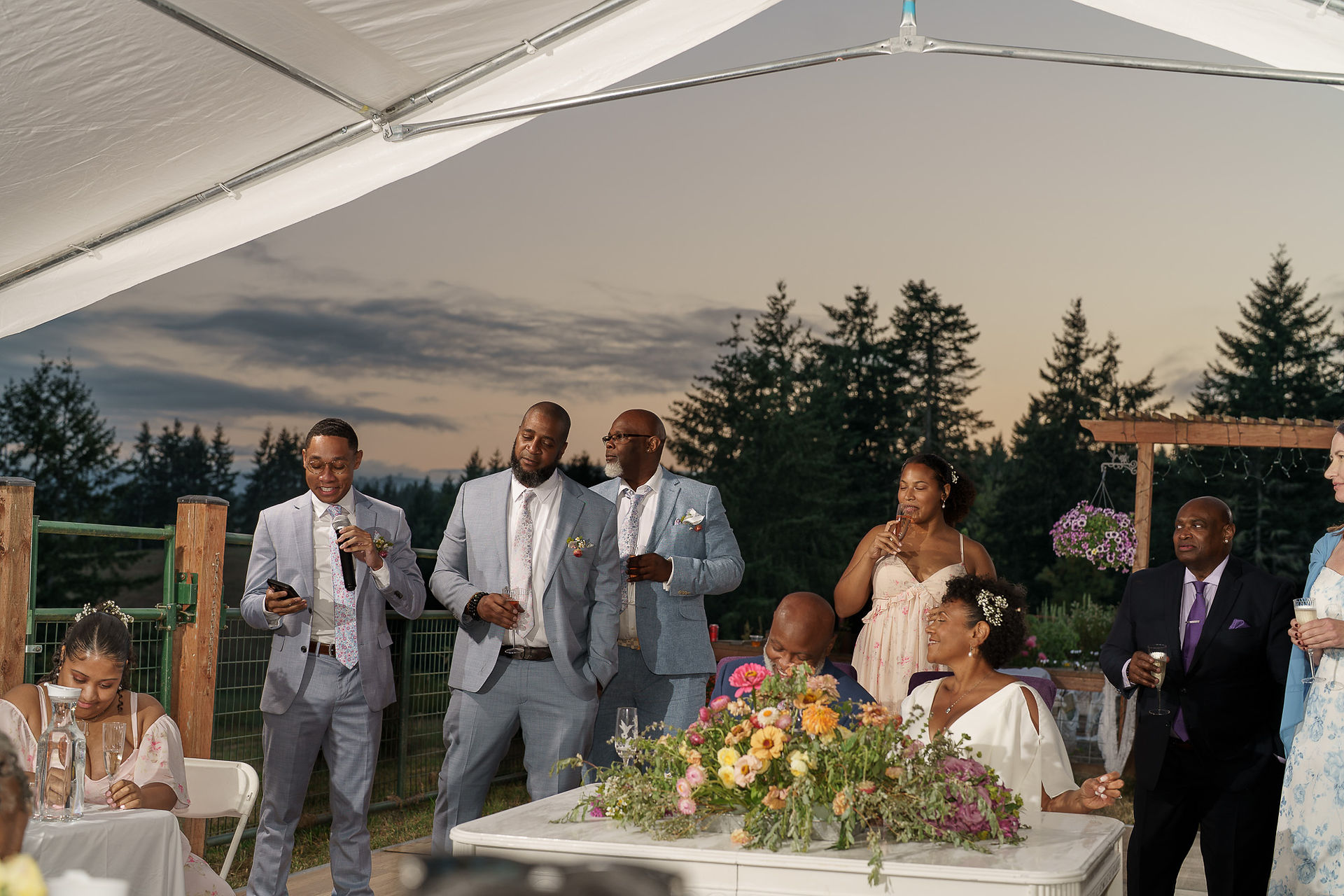 Man in suit speaking into microphone at outdoor farm wedding.