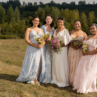 Smiling bride and bridesmaids holding colorful bouquets in a sunny farm field.