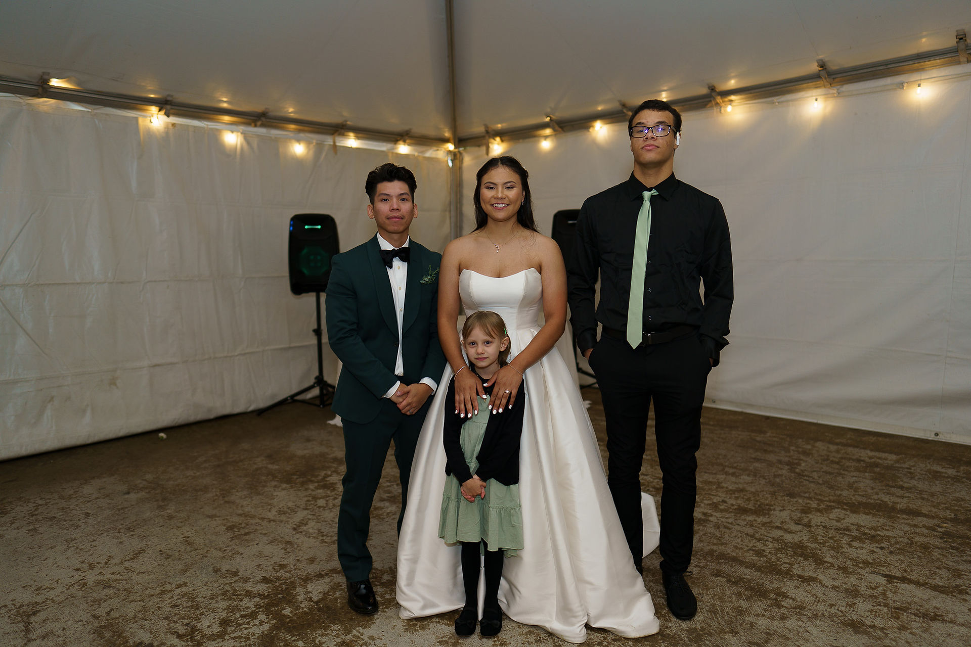 Wedding couple, young girl, and man posing under reception string lights.