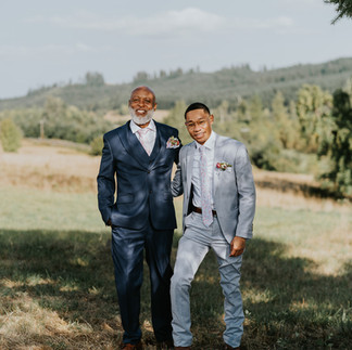 Two men in smart suits smiling outdoors at a farm wedding.