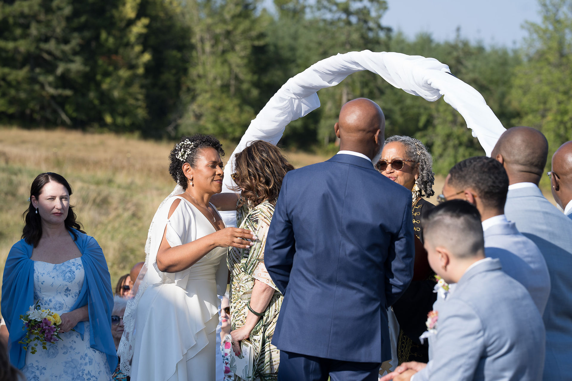 Guests and woman in white dress mingle at outdoor wedding with arch.