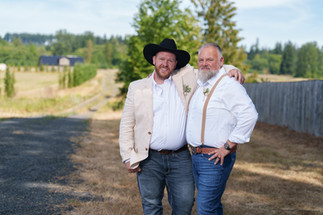 Two smiling grooms, one in cowboy hat, embracing at their summer wedding.
