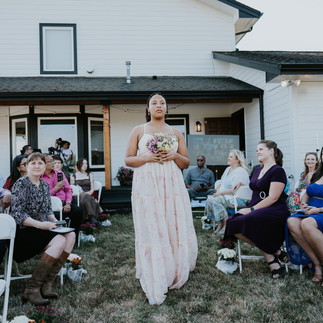Bridesmaid walks down aisle holding flowers at outdoor wedding ceremony.