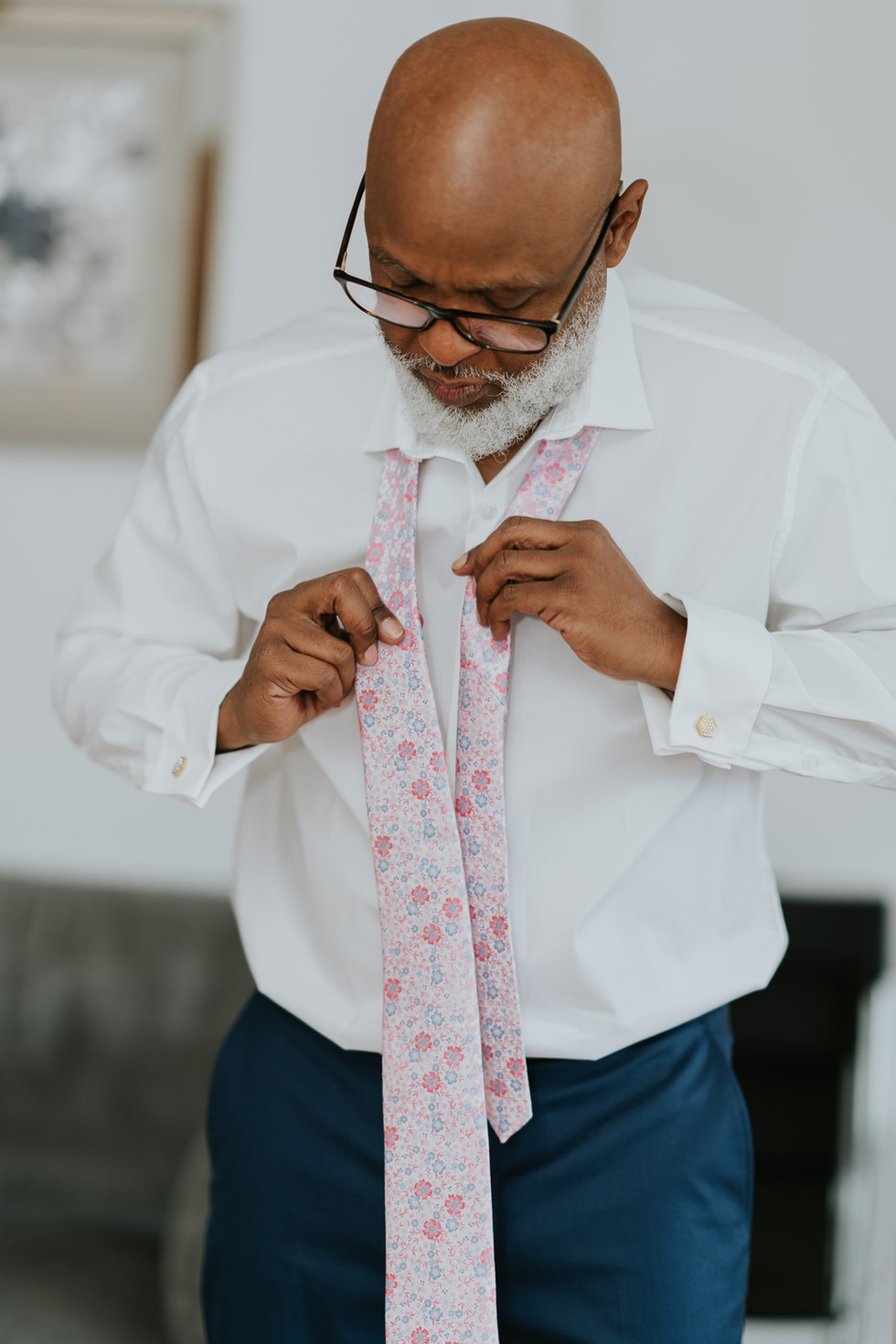 Man with glasses tying a pink floral tie on a white shirt.
