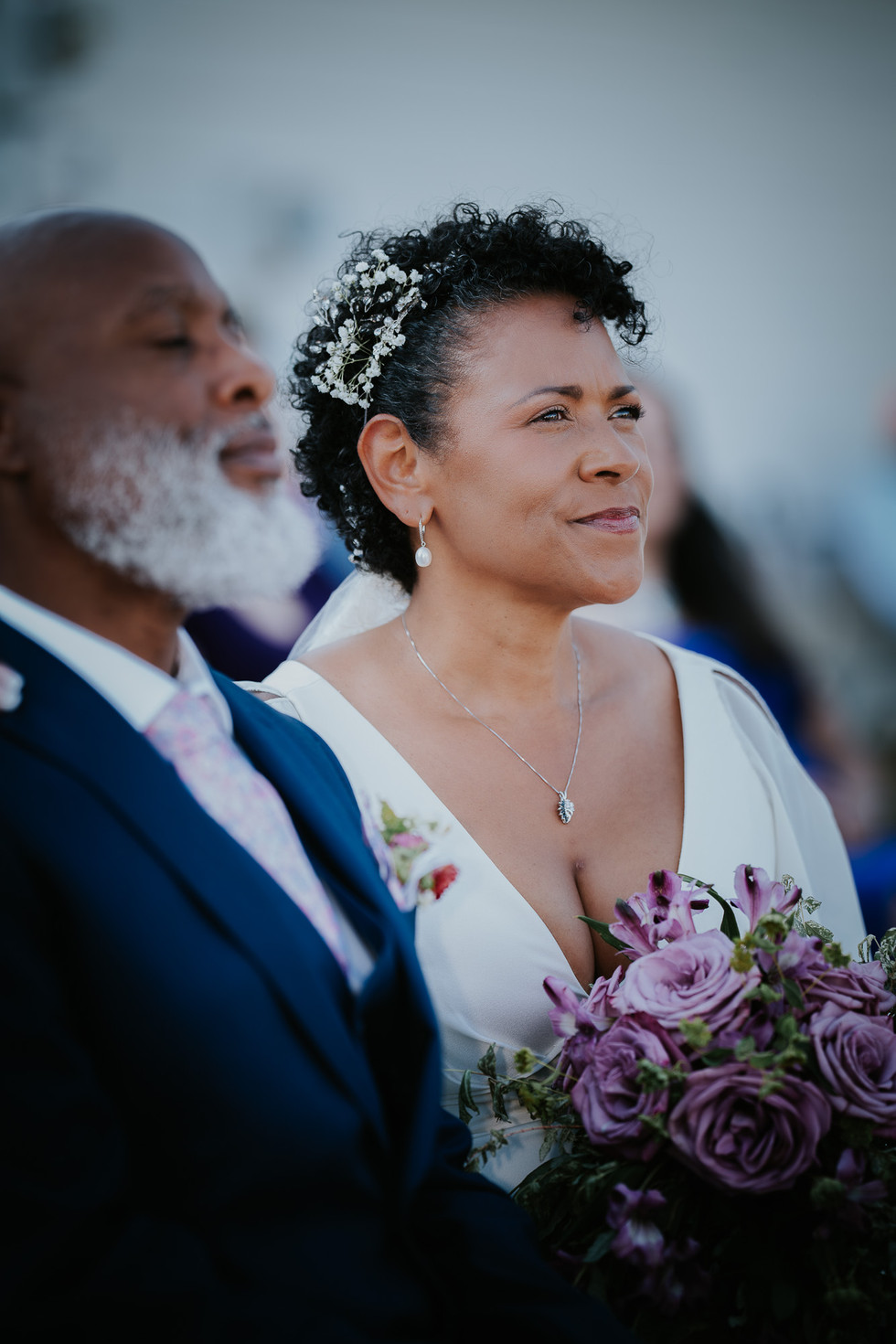 Thoughtful bride in white wedding dress holding purple bouquet beside man.