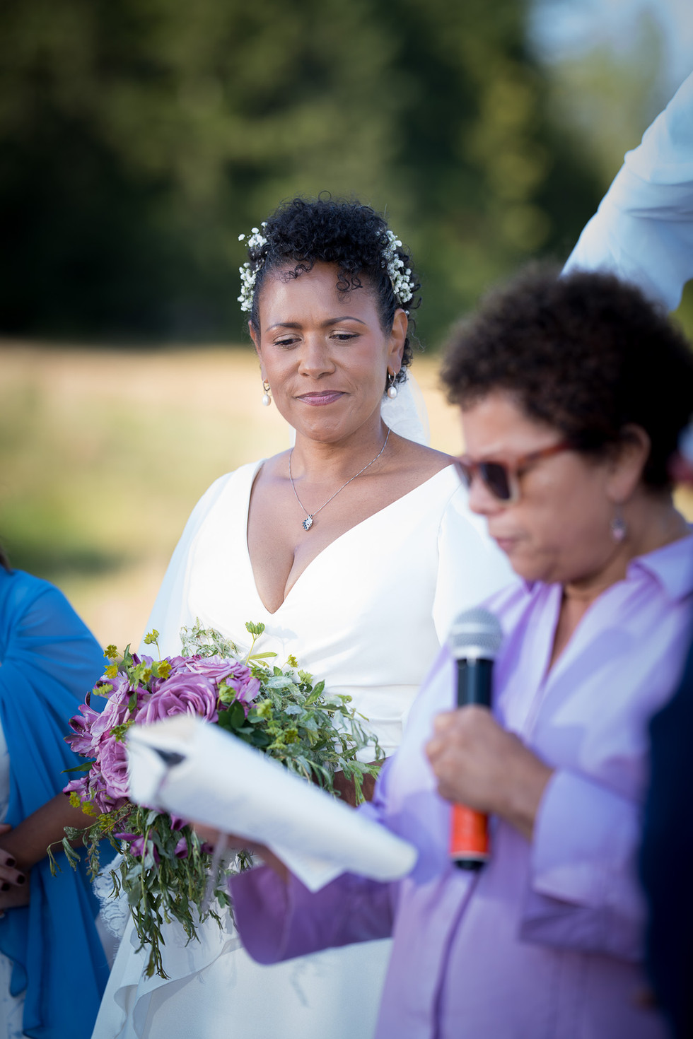 Bride in white dress holding flowers watches speaker intently outdoors.