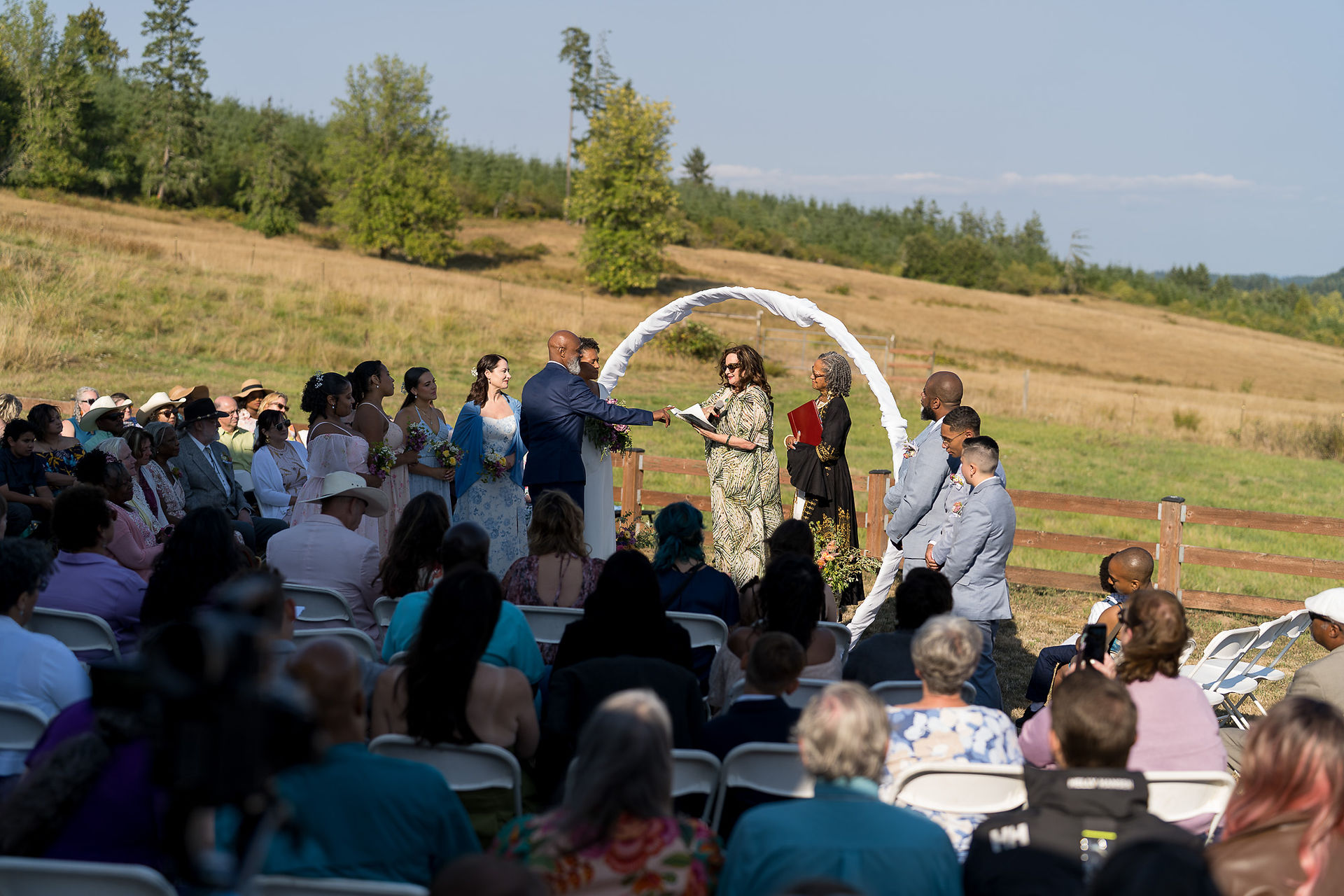 Outdoor wedding ceremony with guests, officiant, and white arch in field.