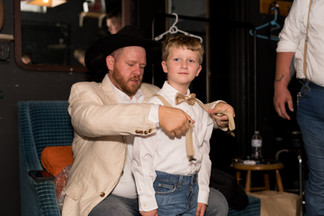Man helps young boy adjust bow tie and suspenders for wedding.
