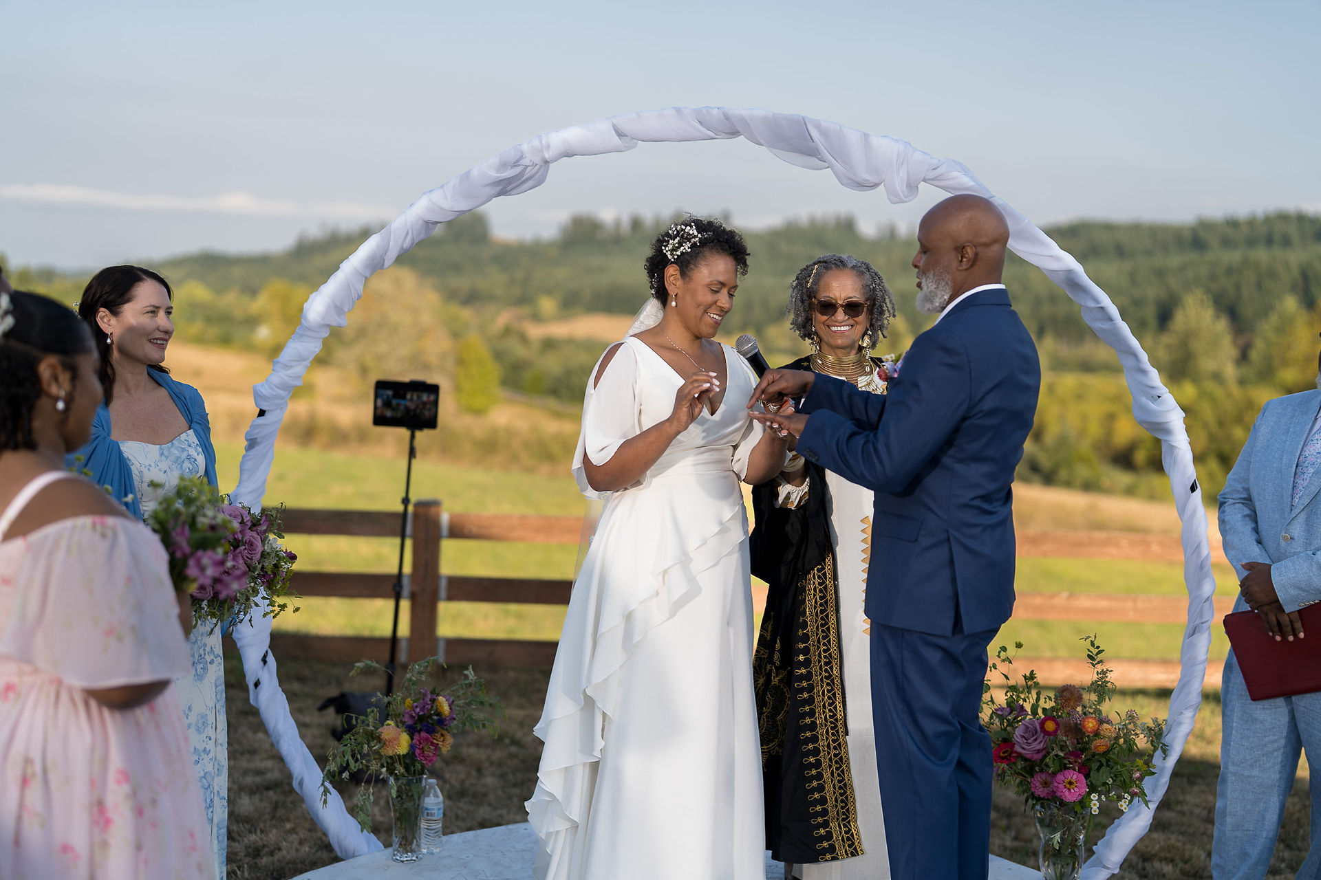 Bride, groom exchanging rings outdoors under white arch with officiant.