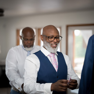 Man in blue vest, pink tie smiles, another man helps prepare.