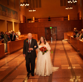 Bride and father walk arm-in-arm down chapel aisle, guests watching.