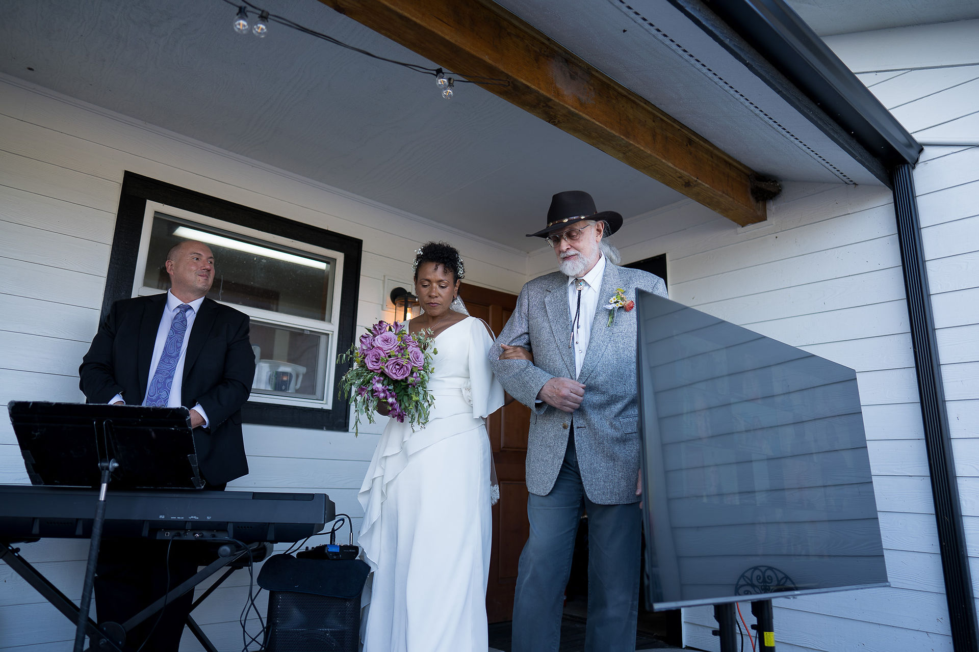 Bride with purple bouquet escorted by older man in hat at wedding.