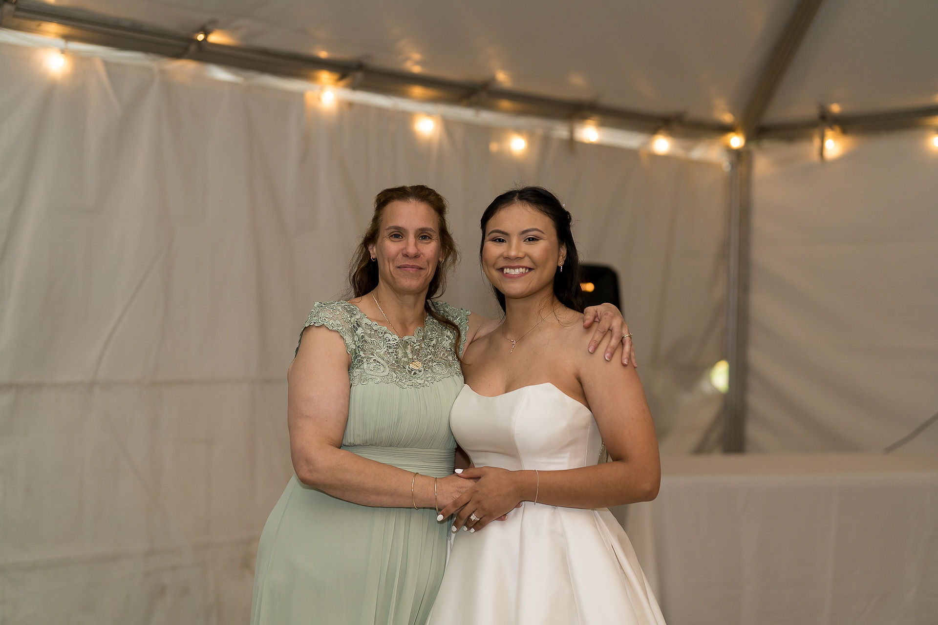 Joyful bride and mother holding hands under festive tent lights.