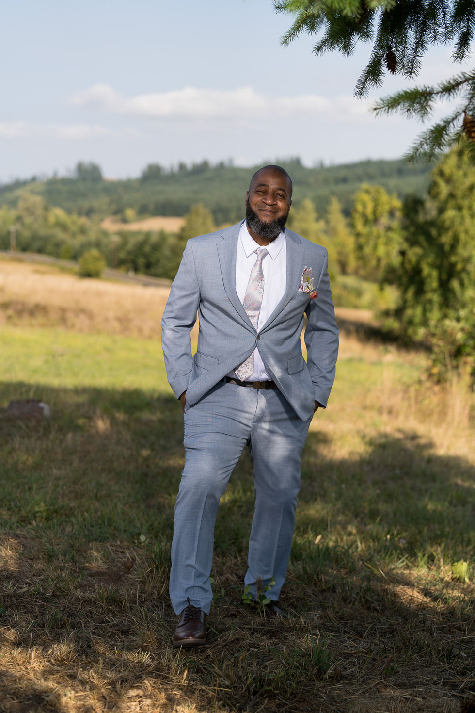 Smiling man in light blue suit standing in grassy field outdoors.