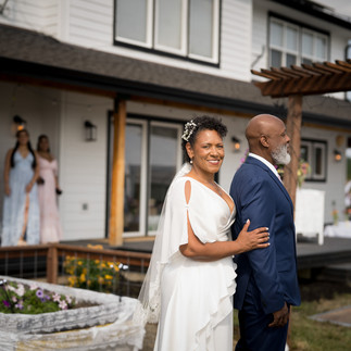 Smiling bride and groom outside white farmhouse with bridesmaids on porch.