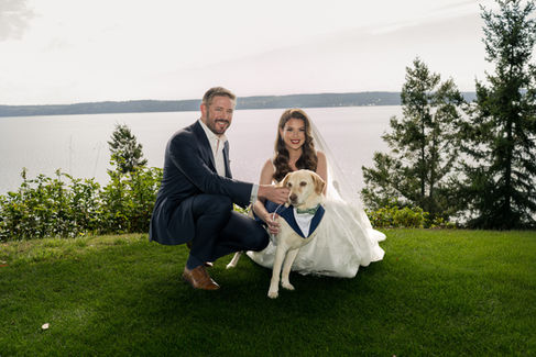 Bride, groom, and yellow lab dog kneeling by water on green grass.