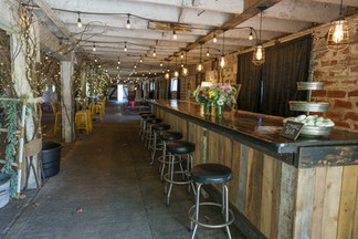 Rustic barn bar with stools, string lights, flowers, and distant seating.