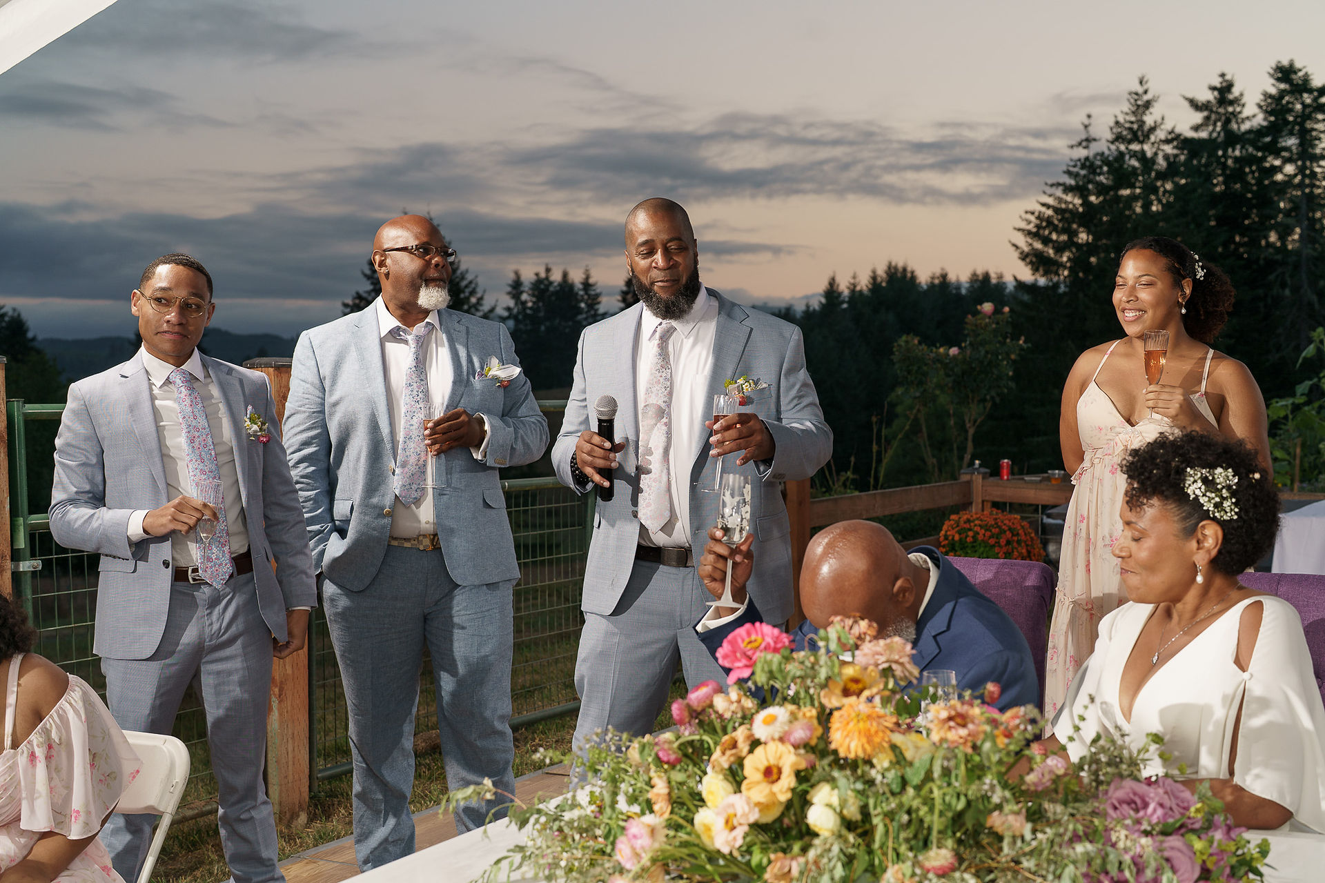 Two groomsmen giving speech, one laughing at intimate farm wedding.