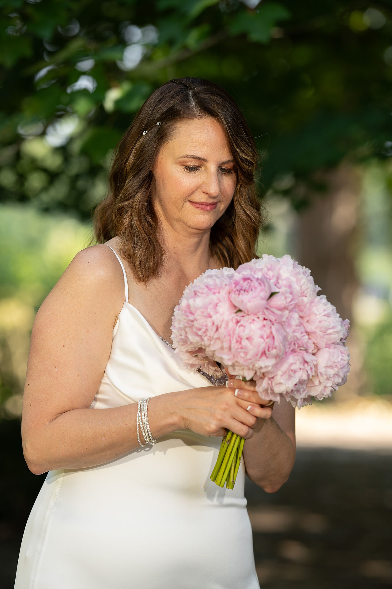 Woman in white dress holding pink peony bouquet outdoors, looking down.