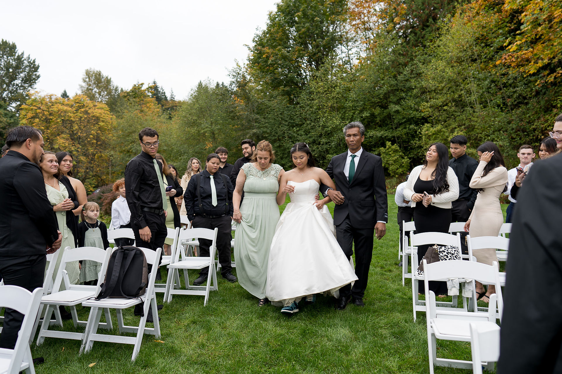 Bride in white gown walking outdoor aisle, escorted by two people.