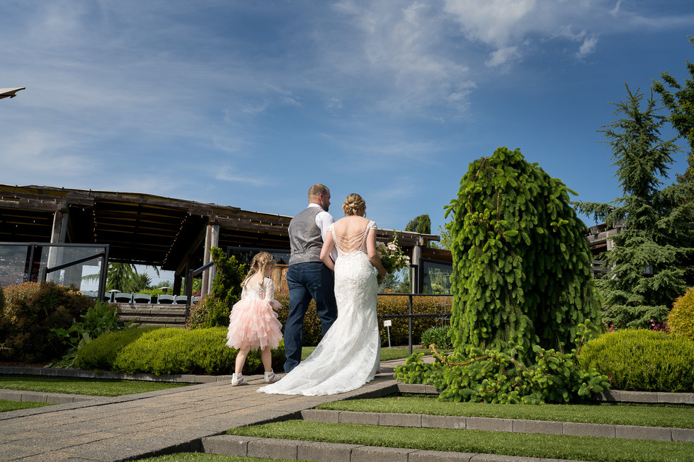 Bride, groom, flower girl walk outdoors on sunny wedding day.