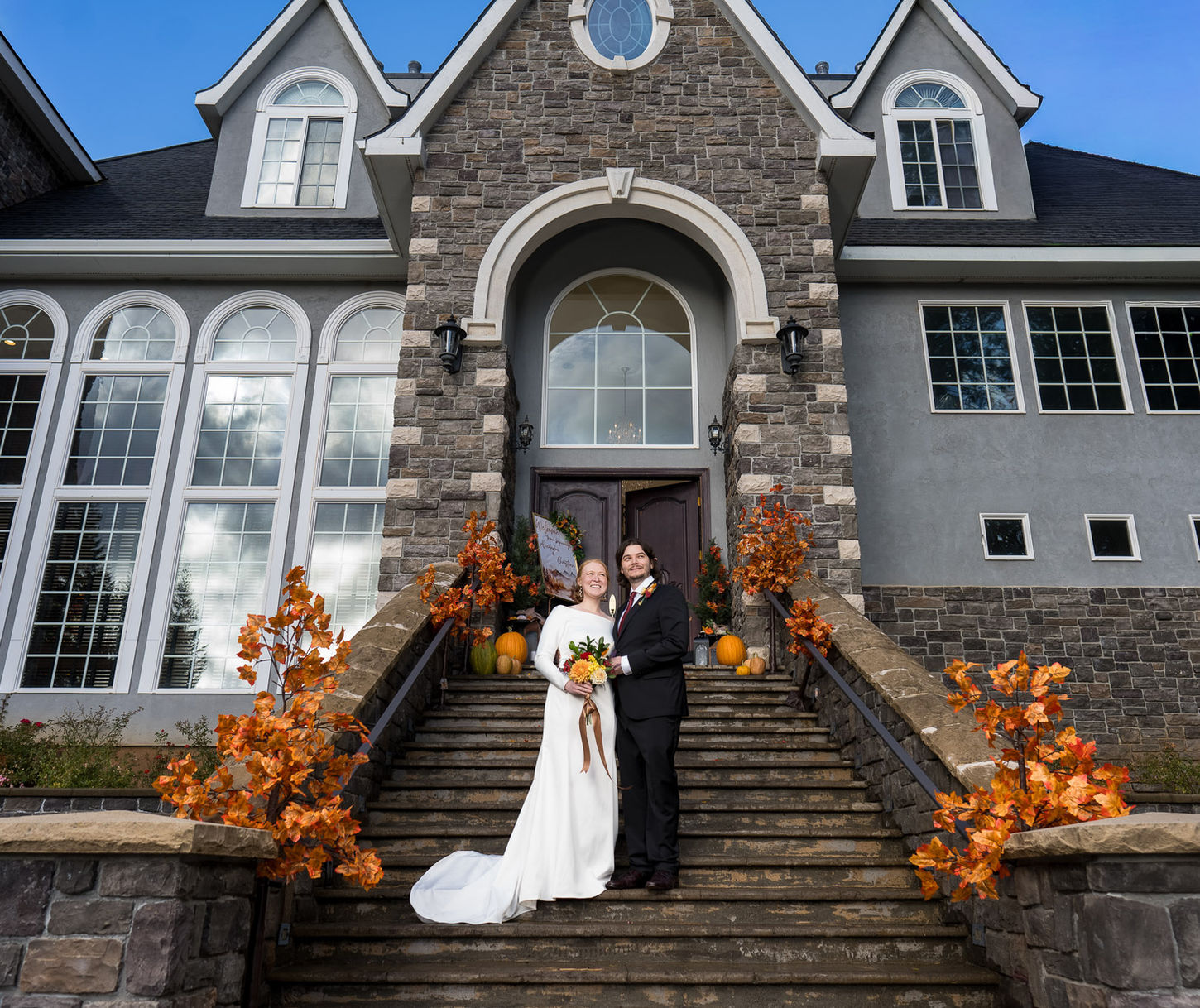 Bride, groom on ornate steps of grand stone house, autumn décor.