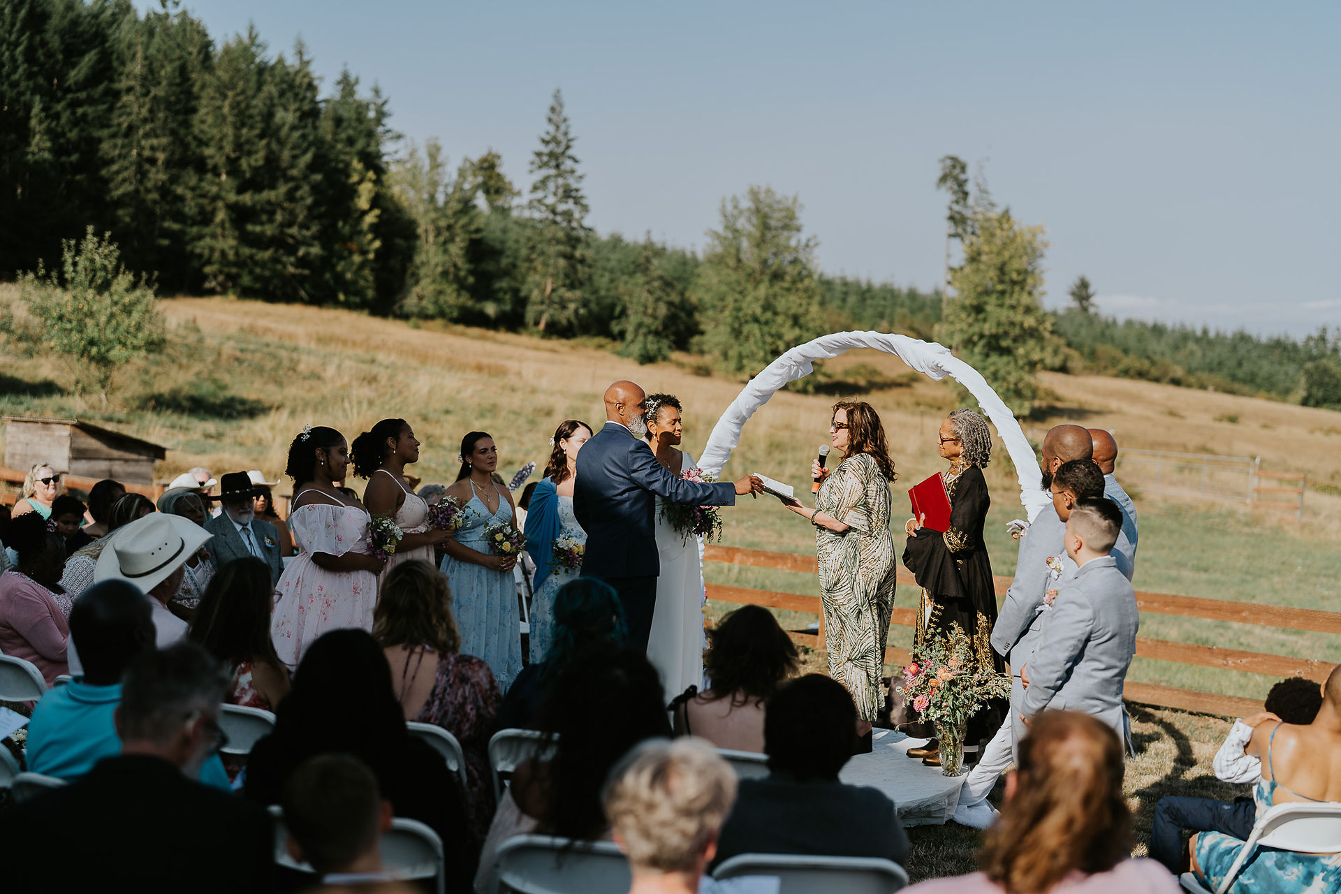 Intimate farm wedding ceremony with couple, officiant, guests, and white arch.