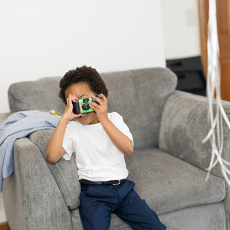 Young boy holding green camera, playing photographer on gray armchair.