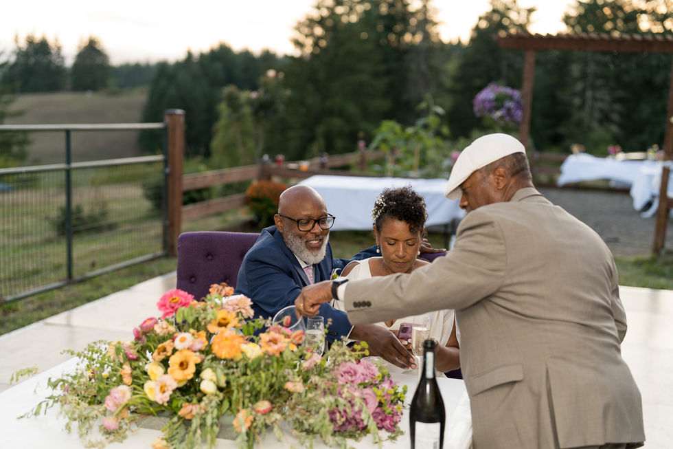 Man pours drinks for couple at outdoor celebration table with flowers.
