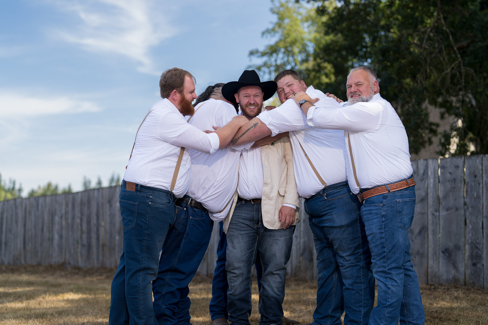 Groom and four groomsmen embracing, laughing outdoors at a wedding.