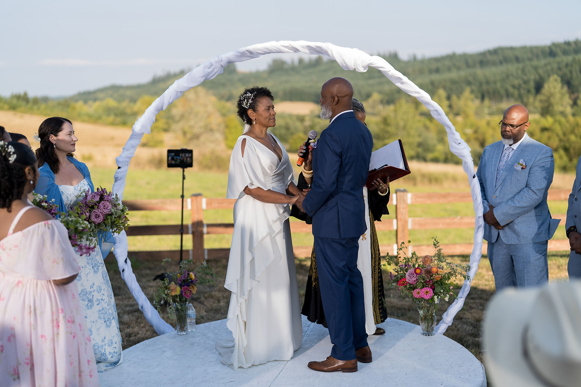 Couple exchanges vows, intimate farm wedding under white arch.