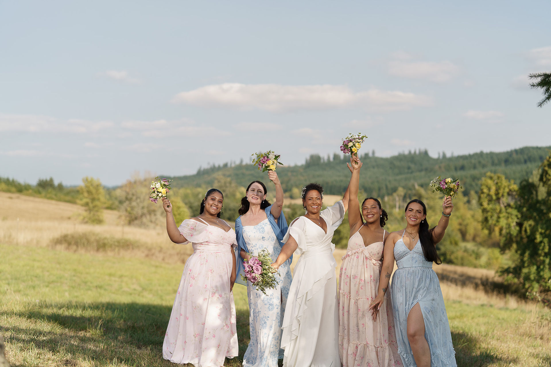 Five women in dresses raising bouquets in a sunny field.