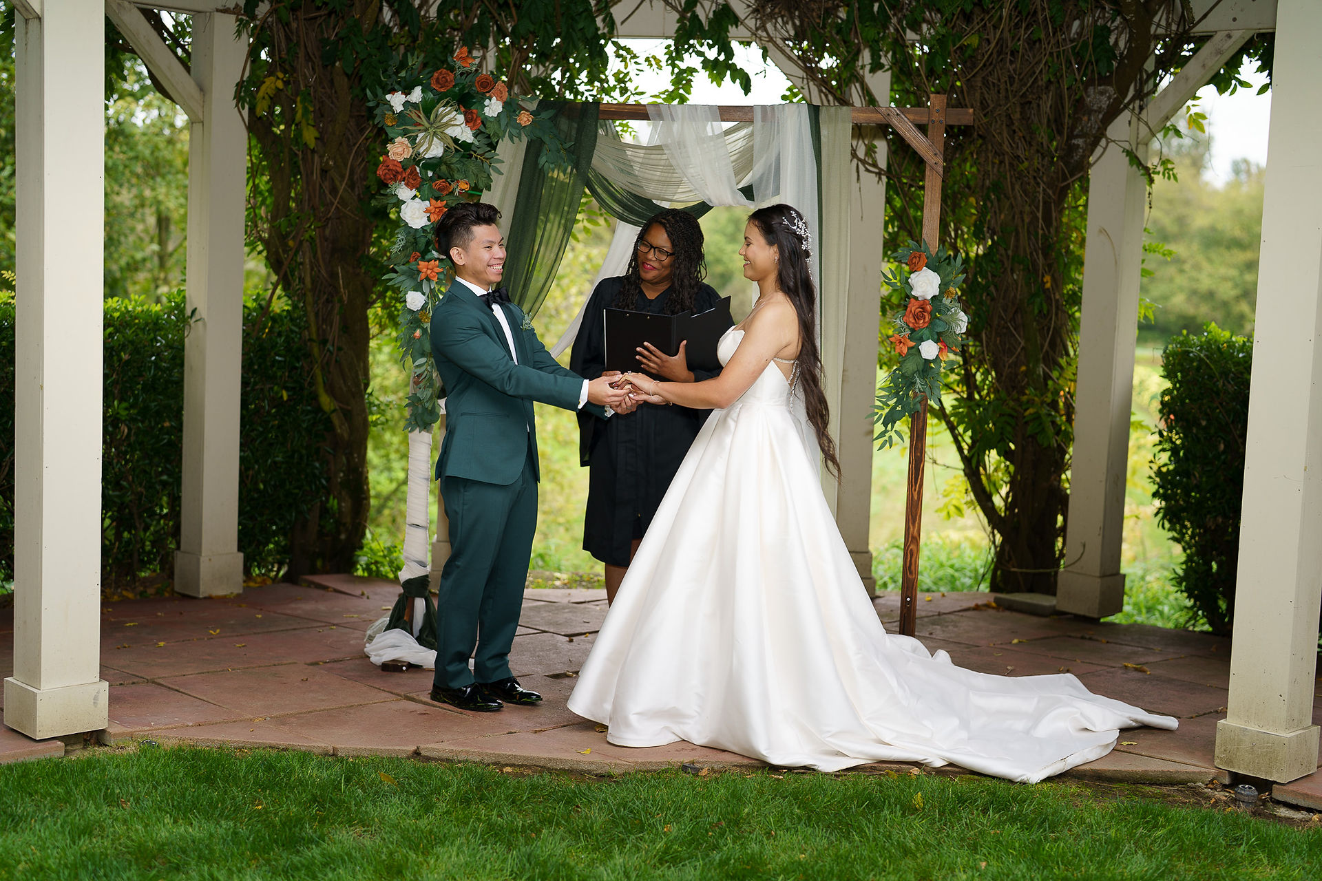 Couple holding hands at outdoor wedding ceremony with officiant and floral arbor.