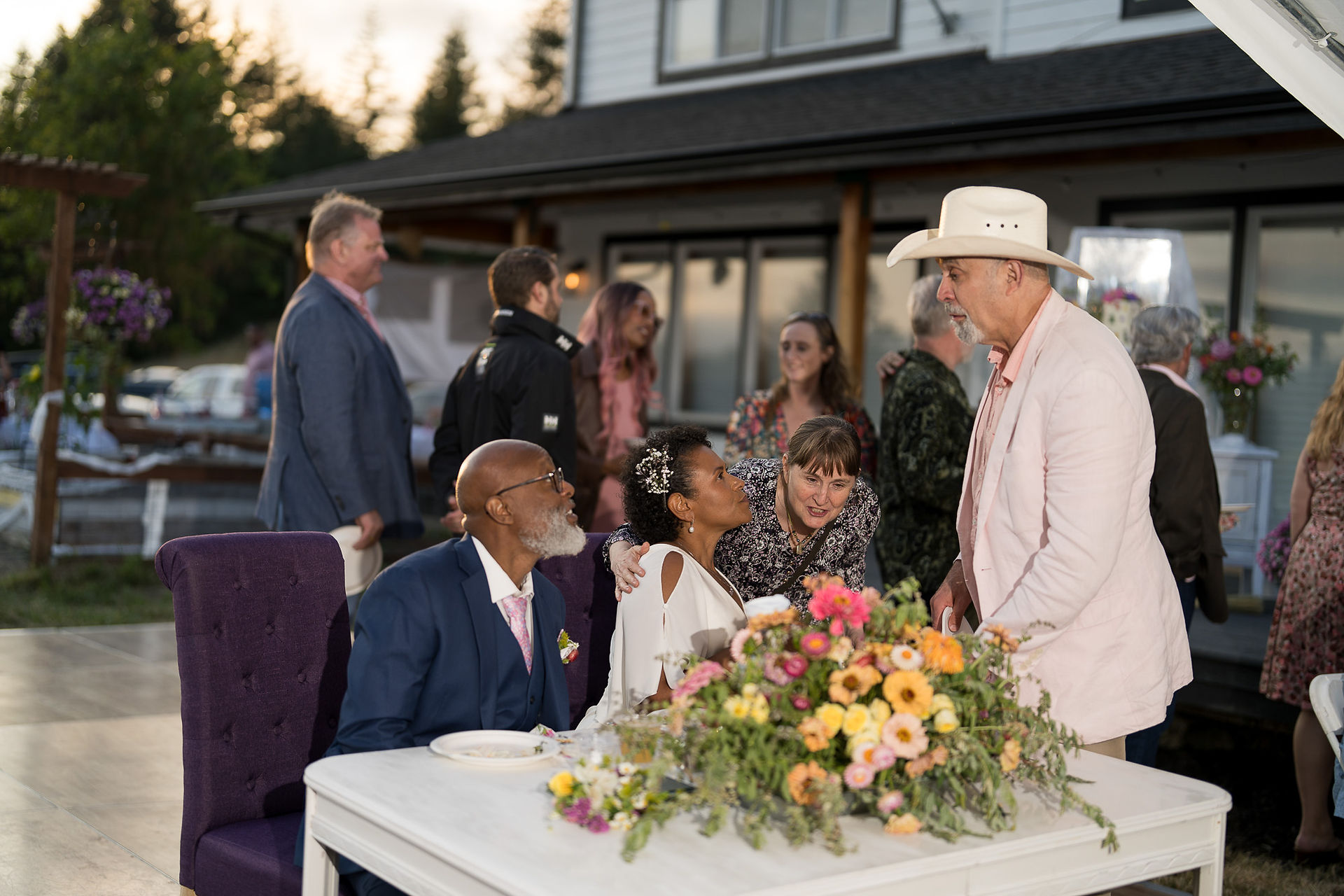 Wedding guests socializing outdoors around table with floral centerpiece.