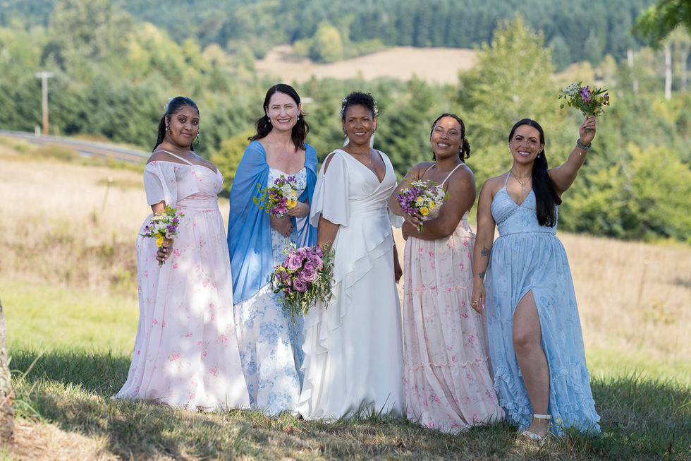 Five smiling women in varied dresses holding bouquets at a farm wedding.